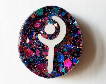 May include: A circular resin badge featuring a dark base filled with multicolored glitter in shades of blue, pink, and purple. A white, stylized graphic symbol resembling a staff or a crescent moon with a central dot is centered on the surface.