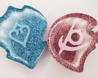 May include: Two resin charms with glittery finishes. The left charm is blue with a white heart-shaped symbol. The right charm is pink with a white circular symbol featuring a curved line. Both items have a smooth, glossy, and semi-translucent appearance.
