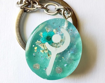 May include: Oval-shaped teal resin keychain with a white symbol and a teal flower. The keychain has silver glitter and dried flowers embedded within the resin. A silver metal chain and keyring are attached.