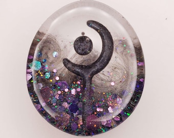 May include: A circular resin cabochon featuring a clear top layer over a dark, swirling background. A black, crescent-shaped symbol sits in the center, surrounded by a dense collection of purple, blue, and iridescent hexagonal glitter flakes.
