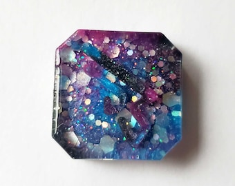 May include: A square-shaped resin cabochon featuring a galaxy-inspired design with deep purple, vibrant blue, and black swirls. The surface is embedded with various sizes of iridescent hexagonal and circular glitter that reflects light.
