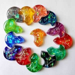 May include: A collection of colorful, resin-cast pendants in various shapes and colors, including green, blue, orange, and red. Each pendant features a constellation design with white lines and glitter accents. The pendants are arranged in a circular pattern.