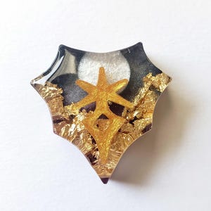 May include: A star-shaped resin pendant with a gold star design. The pendant has a black and white background with gold leaf accents. The edges are clear, creating a layered effect. A decorative accessory.
