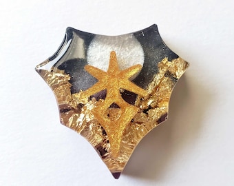May include: A unique, star-shaped resin pendant. The pendant features a gold star design over a black and white background, with gold leaf accents. The edges are clear, giving a layered effect. A decorative accessory.