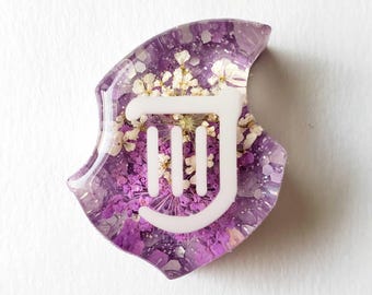 May include: A resin-cast shield-shaped item with a white symbol and dried flowers in shades of purple and white. The symbol is a stylized design with three vertical lines and a curved top.