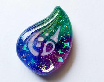 May include: A teardrop-shaped resin pendant featuring a gradient of green, blue, and purple colors with embedded holographic glitter. The surface displays a white crescent moon symbol, several small star shapes, and a metallic gold crescent moon charm.