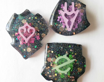 May include: Three resin-cast charms with glitter and floral inclusions. Each charm features a different symbol in pink or green. The shapes are shield-like, with a dark background and glitter.