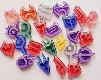 May include: A collection of small, colorful resin charms in various shapes, including shields, stars, and abstract symbols. Each piece features a translucent border and a glitter-filled center in shades of red, blue, purple, green, orange, and yellow.