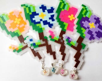 May include: Four pixel art flower charms made from fused plastic beads. Each flower features a brown stem, green leaves, and a distinct petal color: orange, blue, pink, and purple. Each stem is attached to a small, clear, heart-shaped resin charm.