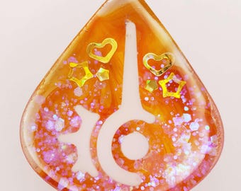 May include: A teardrop-shaped resin charm featuring a vibrant orange swirled background embedded with iridescent pink glitter, gold-colored heart and star-shaped sequins, and a prominent white graphic symbol resembling a stylized sun or gear.