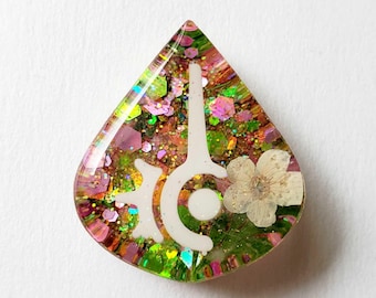 May include: A teardrop-shaped resin pendant featuring a white stylized symbol, a pressed white flower, and a dense mixture of iridescent pink, green, and gold hexagonal glitter embedded within the clear, glossy, and smooth material.