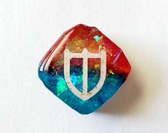 May include: A colorful, diamond-shaped resin piece with a white shield design. The top half is red, transitioning to blue at the bottom. Glitter adds sparkle. A unique, handcrafted accessory.