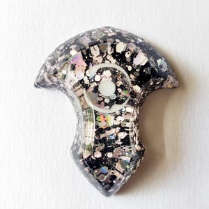 May include: A decorative resin pendant with a black base, speckled with silver and iridescent glitter. The pendant features a white, eye-shaped design in the center. The overall shape is a stylized, rounded form with a pointed bottom.