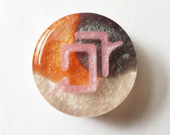 May include: A circular resin cabochon featuring a swirling blend of metallic orange, dark charcoal, and pearlescent white colors. The surface is embedded with a raised, light pink symbol that resembles two connected, stylized geometric shapes.