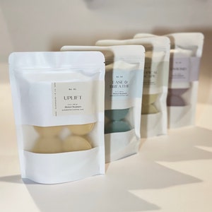 May include: Four white pouches with clear windows, each containing shower steamers. The first pouch reads "Uplift" and the second reads "Ease & Breathe". The steamers are various pastel colors, including yellow, green, and purple. The packaging is minimalist.