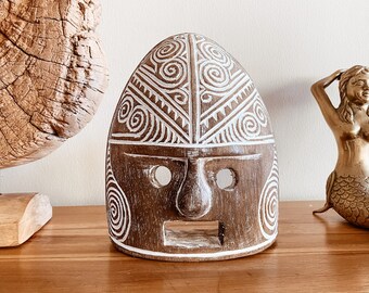 Puede incluir: Una máscara de madera con diseños geométricos pintados en blanco. La máscara tiene dos agujeros para los ojos y una abertura para la boca. La máscara está tallada en estilo tribal.