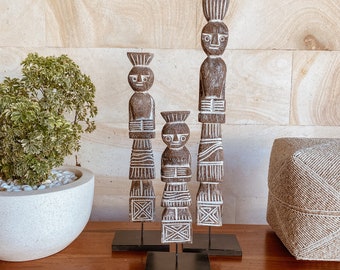 Puede incluir: Tres figuritas de madera con detalles pintados en blanco se colocan sobre bases negras. Las figuritas están talladas en forma de personas con rasgos estilizados y patrones geométricos. Las figuritas están dispuestas en fila sobre una superficie de madera.