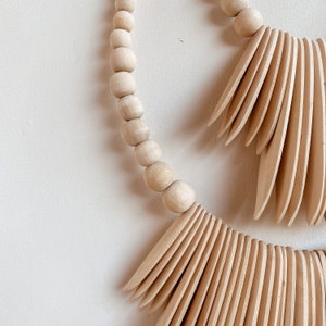 May include: A wooden wall hanging with a beaded circle and two fan-shaped wooden pieces. The beads are natural wood and the fan pieces are a light brown color.