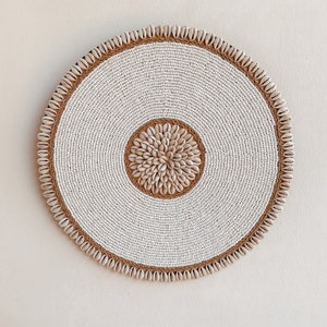 Op de afbeelding: Een ronde wanddecoratie gemaakt van witte kralen en natuurlijke schelpen. Het midden van de decoratie heeft een cirkelvormig ontwerp van schelpen in een bruine tint.