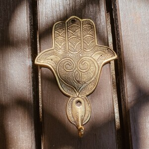 Könnte beinhalten: Ein goldfarbener Hamsa-Hand-Wandhaken aus Metall mit filigranen Details und einem Haken unten.