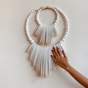May include: White beaded wall hanging with two concentric circles and a fringe of white tassels.