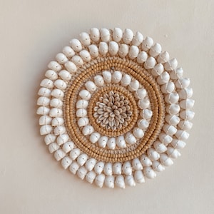 May include: A circular wall decor made of white seashells and natural brown beads arranged in a concentric pattern. The center of the decor features a smaller circle of brown seashells.