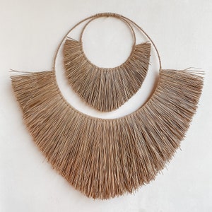 May include: A large, brown, woven wall hanging with a fringe design. The hanging is made of natural fibres and has a circular shape with two concentric circles.