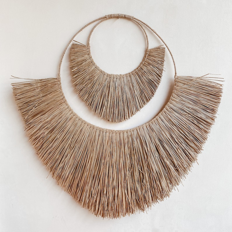 May include: A large, brown, woven wall hanging with a fringe design. The hanging is made of natural fibres and has a circular shape with two concentric circles.
