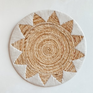 May include: A round, woven placemat with a sunburst design. The center is made of natural-colored straw, with a white, triangular-shaped border. The placemat is designed to protect a table from heat and spills.