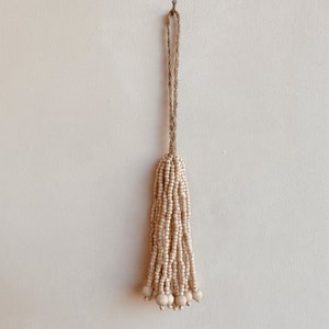May include: A white beaded tassel with a natural fiber cord hanging from a hook. The tassel is made of small, round beads and has a fringe at the bottom.