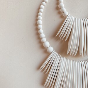 May include: White wooden bead garland with two white wooden fringe tassels. The garland is in the shape of a circle and the tassels are hanging down.