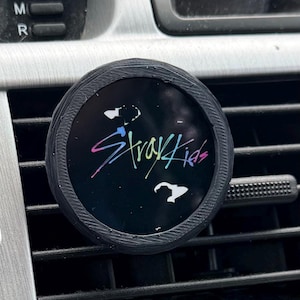 May include: A black car air vent clip featuring the text "Stray Kids" in a colorful, stylized font. The clip is round with a textured edge and is set against a silver and black car dashboard.