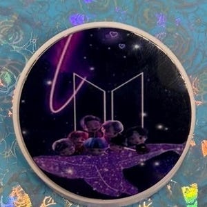 May include: A round, white pop socket featuring a design with a dark purple and black background. The design includes the letters "M" and "B" in white, with small cartoon figures and a purple shooting star.