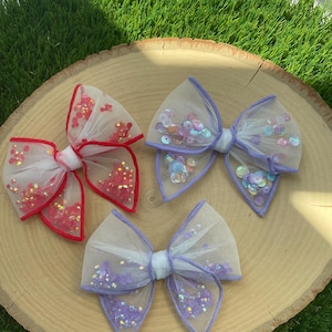 May include: Three hair bows made of white tulle with different colored sequins. The bows are arranged on a wooden surface.