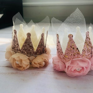 May include: Two miniature crowns with pink and gold glitter, adorned with tulle and delicate pink and beige fabric flowers. Each crown has a pearl accent in the center of each flower.
