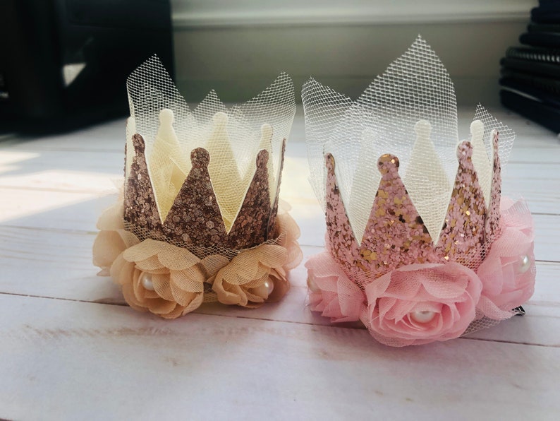 May include: Two miniature crowns with pink and gold glitter, adorned with tulle and delicate pink and beige fabric flowers. Each crown has a pearl accent in the center of each flower.