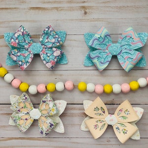 May include: Four handmade hair bows with different patterns. The bows are made of fabric with glitter and have a white backing. The bows are arranged in two rows of two.