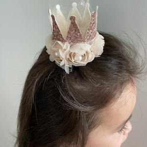 May include: A small, glittery crown with a tulle base and a floral accent. The crown is rose gold and the flowers are ivory.