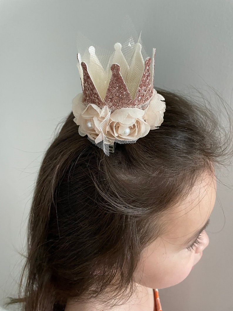 May include: A small, glittery crown with a tulle base and a floral accent. The crown is rose gold and the flowers are ivory.