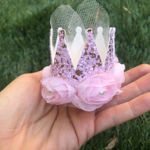 May include: A pink and gold glitter crown with a tulle veil and pink fabric flowers with pearl centers.