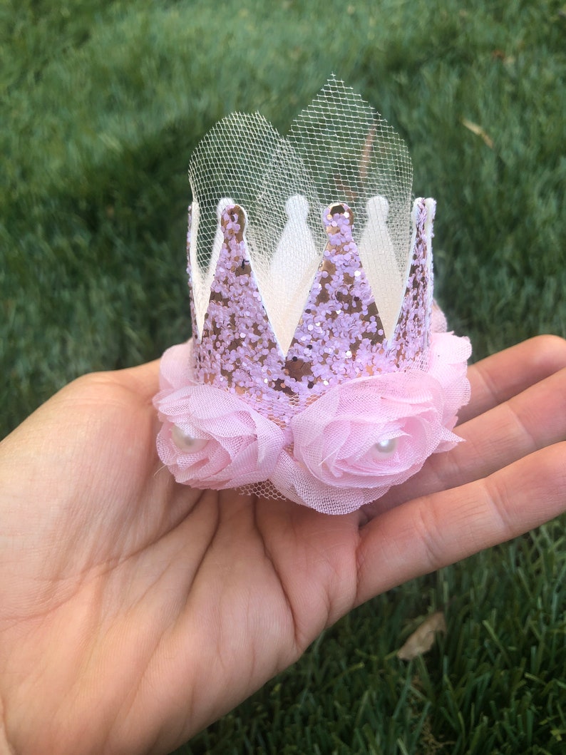 May include: A pink and gold glitter crown with a tulle veil and pink fabric flowers with pearl centers.