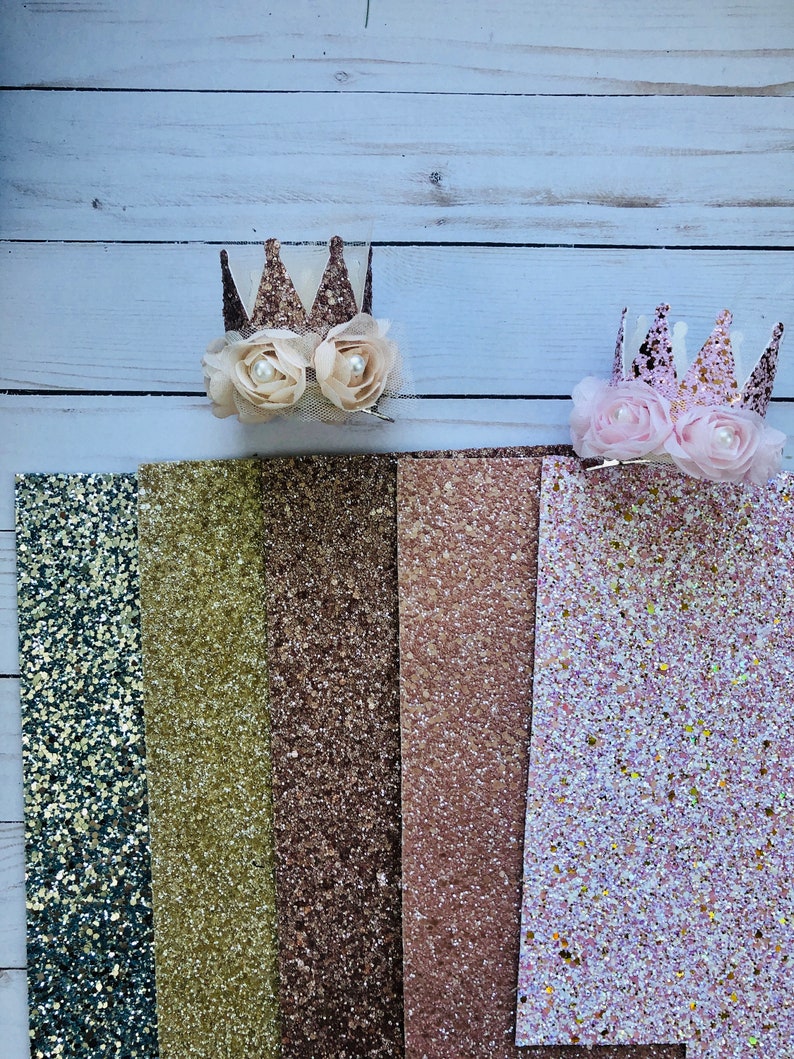 May include: Six strips of glitter fabric in various colors, including teal, gold, brown, rose gold, and pink. Two glitter crowns with white and pink flowers are on top of the fabric.