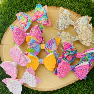 May include: Assortment of colorful hair bows with various patterns and textures. The bows are made of glitter, fabric, and floral prints. They are arranged on a wooden surface.