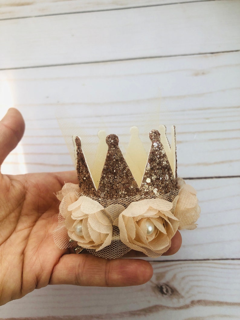 May include: A small gold glitter crown with a tulle base and two beige fabric flowers with pearl centers.