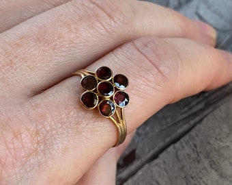 Puede incluir: Un anillo de oro con un grupo de siete piedras preciosas redondas de color rojo oscuro engastadas en forma de flor.
