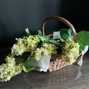 May include: A wicker basket filled with a bouquet of light green hydrangeas. The basket has a handle and is sitting on a dark surface.