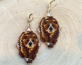 May include: A pair of handmade beaded earrings in a diamond shape. The earrings feature a geometric pattern in shades of brown, tan, and white, with a gold-colored hook closure. The earrings are displayed on a wooden surface.