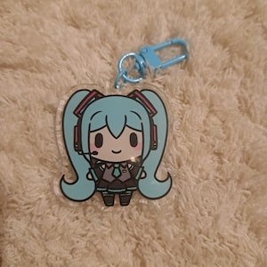 May include: An acrylic keyring featuring a chibi-style character with long teal twin-tails, wearing a dark grey outfit with a teal tie and red-accented headphones. The character has a small smile and is attached to a light blue metal lobster clasp.