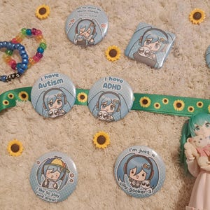 May include: A collection of light blue and white pinback buttons with anime-style illustrations and text, including "I have Autism" and "I have ADHD." Also, a green ribbon with sunflower designs, a rainbow beaded bracelet, and a figurine.