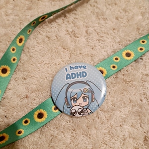May include: A light blue button with the text "I have ADHD" and a cartoon character illustration. The button is on a green lanyard decorated with yellow sunflowers. The lanyard is on a white, textured surface.